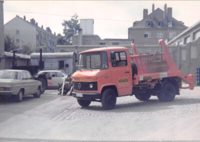 Orangefarbener HAAF Containerdienst-LKW auf einem Platz mit Gebäuden im Hintergrund.