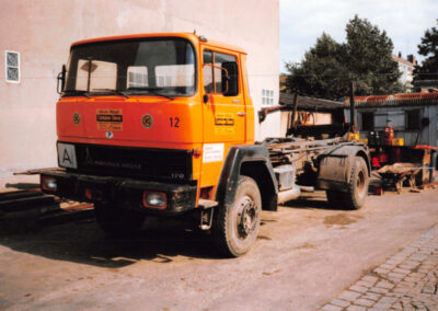 Orangefarbener LKW ohne Aufbau steht neben einem Gebäude auf einem Hof.