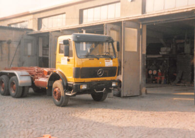 Gelb‑orangefarbener LKW steht vor einer geöffneten Halle mit gepflastertem Hof.