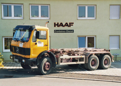 Gelb-oranger LKW ohne Aufbau vor der beschrifteten Fassade „HAAF Container-Dienst“.