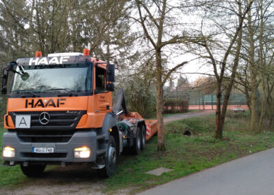 Orangefarbener HAAF-Sattelzug fährt auf einer schmalen Straße an Bäumen vorbei.
