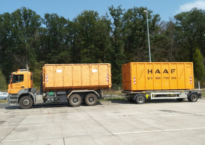 LKW mit Anhänger und zwei gelben HAAF-Containern steht auf einem asphaltierten Platz vor Bäumen.