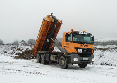 Orangefarbener HAAF-LKW kippt seine Mulde auf einer verschneiten Fläche ab, im Hintergrund Holz- oder Erdhaufen.