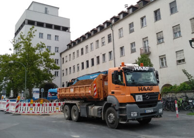 Orangefarbener HAAF-LKW fährt an einer Baustelle in einer Innenstadtstraße mit Absperrgittern und mehrstöckigen Häusern vorbei.