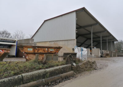 Offene Halle mit Wellblechdach, davor Container und gelagerte Materialien auf dem Betriebsgelände.