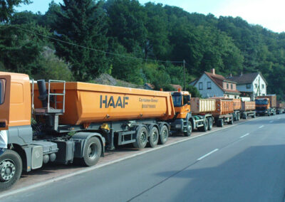 Mehrere orangefarbene HAAF-LKW fahren hintereinander durch eine Ortschaft, rechts Häuser, links Grünflächen.