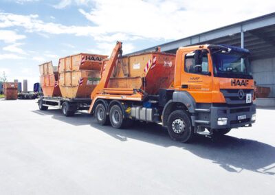 Mehrere orangefarbene HAAF-Container-LKW stehen hintereinander auf einem Hof vor einer Halle.