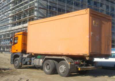 Orangefarbener HAAF-Container auf einem LKW-Auflieger vor einem eingerüsteten Gebäude auf einer Baustelle.
