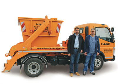 Kleiner orangefarbener HAAF-LKW mit Absetzcontainer, daneben stehen Markus Haaf und Alexander Haaf.
