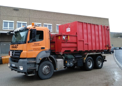 Orangefarbener HAAF-LKW mit rotem Großcontainer steht vor einer Industriehalle.