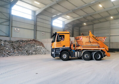 Große, helle Halle mit Betonboden, seitlichen Schüttgut-Haufen und einem orangefarbenen HAAF-LKW im Inneren.
