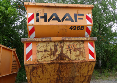 Mehrere orangefarbene HAAF-Container stehen an einer Hallenwand auf einem befestigten Hof, im Hintergrund Bäume.