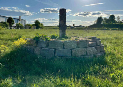 Alte runde Steinmauer oder Brunnenrest steht auf einer Wiese, im Hintergrund Felder und Himmel mit Wolken.
