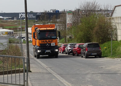 Orangefarbener HAAF-LKW mit Container fährt durch ein Gewerbe- bzw. Mischgebiet, im Hintergrund Gebäude und Autos.