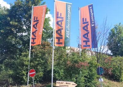 Drei hohe Fahnenmasten mit HAAF-Flaggen vor Bäumen und blauem Himmel.