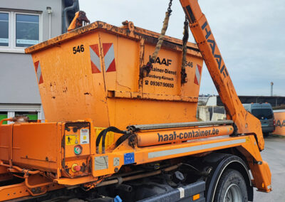 Orangefarbener HAAF-Container hängt am Haken eines LKW-Krans, seitliche Detailansicht.