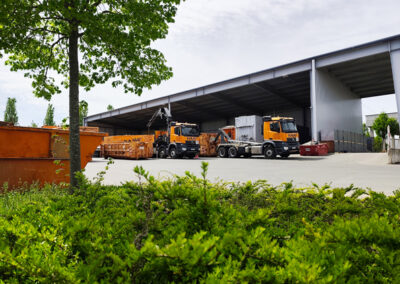 HAAF-Container und Baumaschinen stehen vor einer großen offenen Halle, im Vordergrund grüne Büsche.