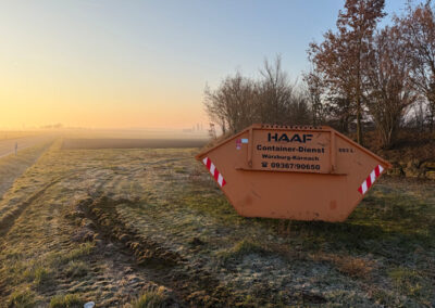 Orangefarbener HAAF-Container steht auf einer frostigen Wiese im Morgenlicht, im Hintergrund Bäume und Felder.