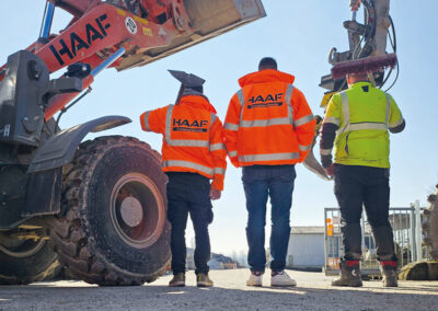 Drei Personen in orangefarbener HAAF-Warnkleidung und Helm stehen auf dem RecyclingCenter-Gelände neben einem großen Radlader.